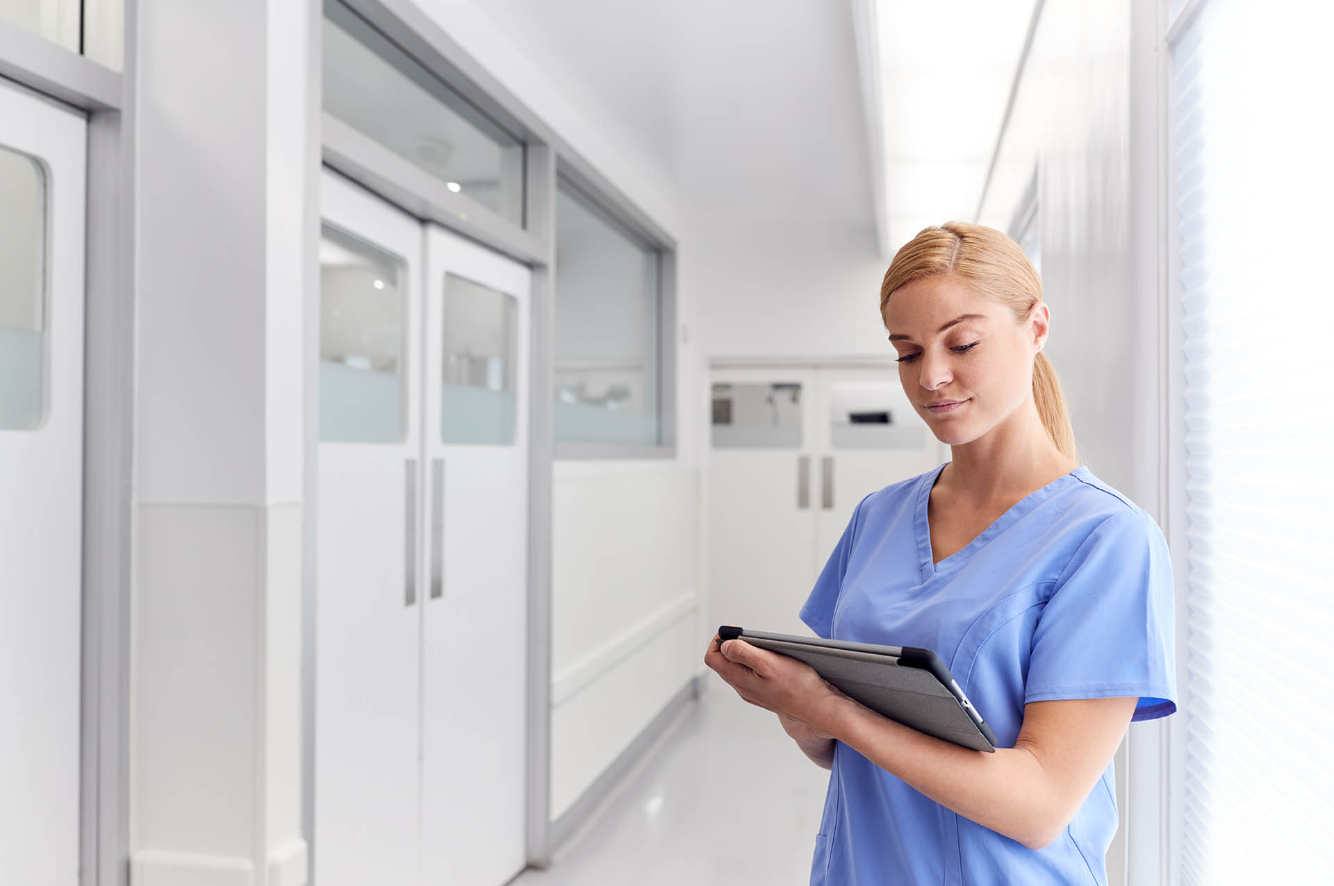 Female Doctor Wearing Scrubs In Hospital Corridor Using Digital Tablet